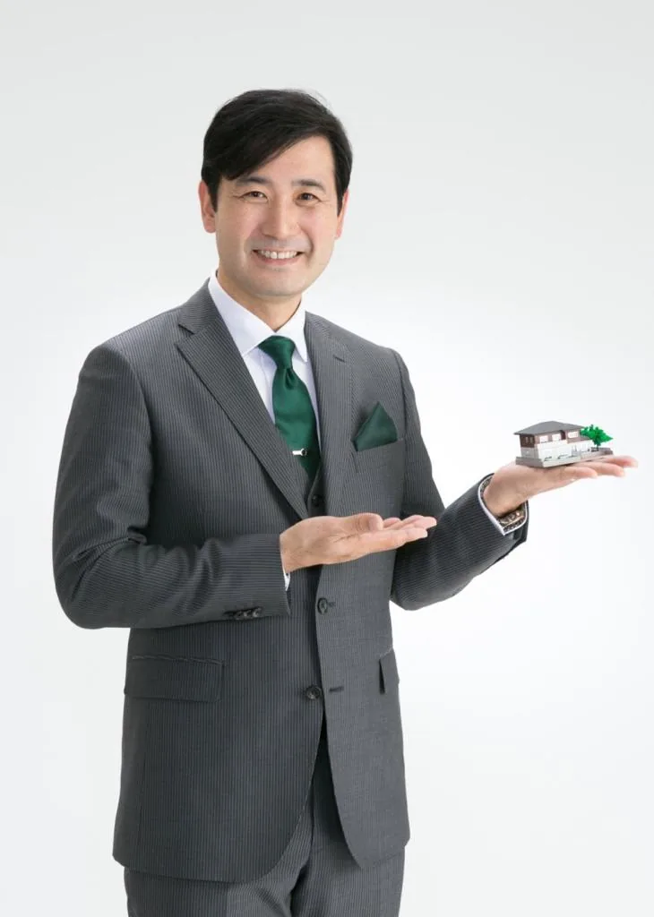 Koji Muto, fundador e diretor-presidente da Ji Consulting, de terno e gravata verde, segurando uma maquete de casa